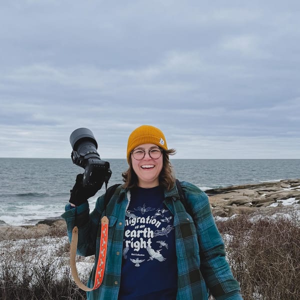 Stephanie smiles on a rocky winter coastline, holding up a camera with a long lens. She wears a yellow beanie, plaid flannel, and a Bird Collective t-shirt.